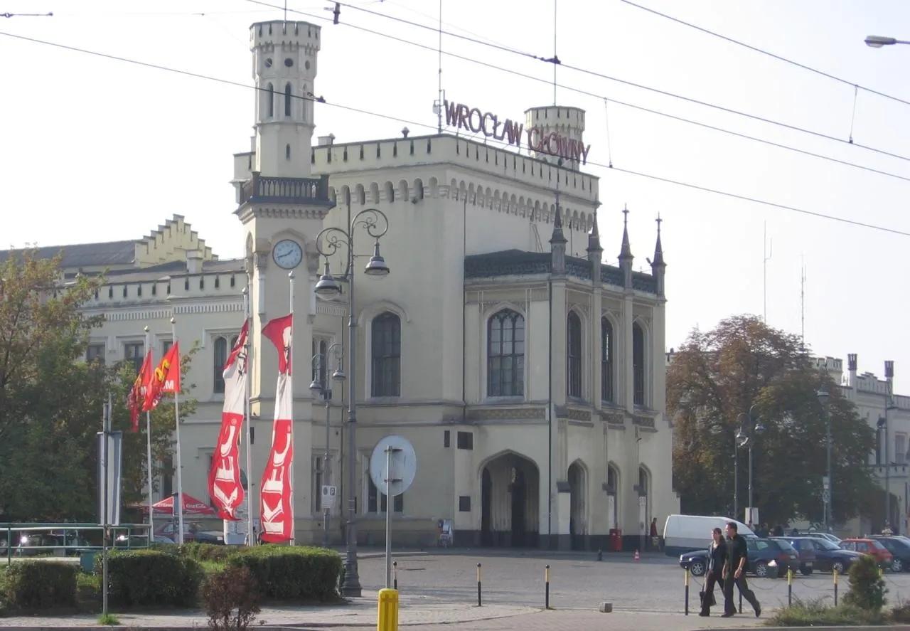 Szaro-kremowa fasada Dworca Głównego przed remontem; przed wejściem flagi reklamowe i zaparkowane samochody.