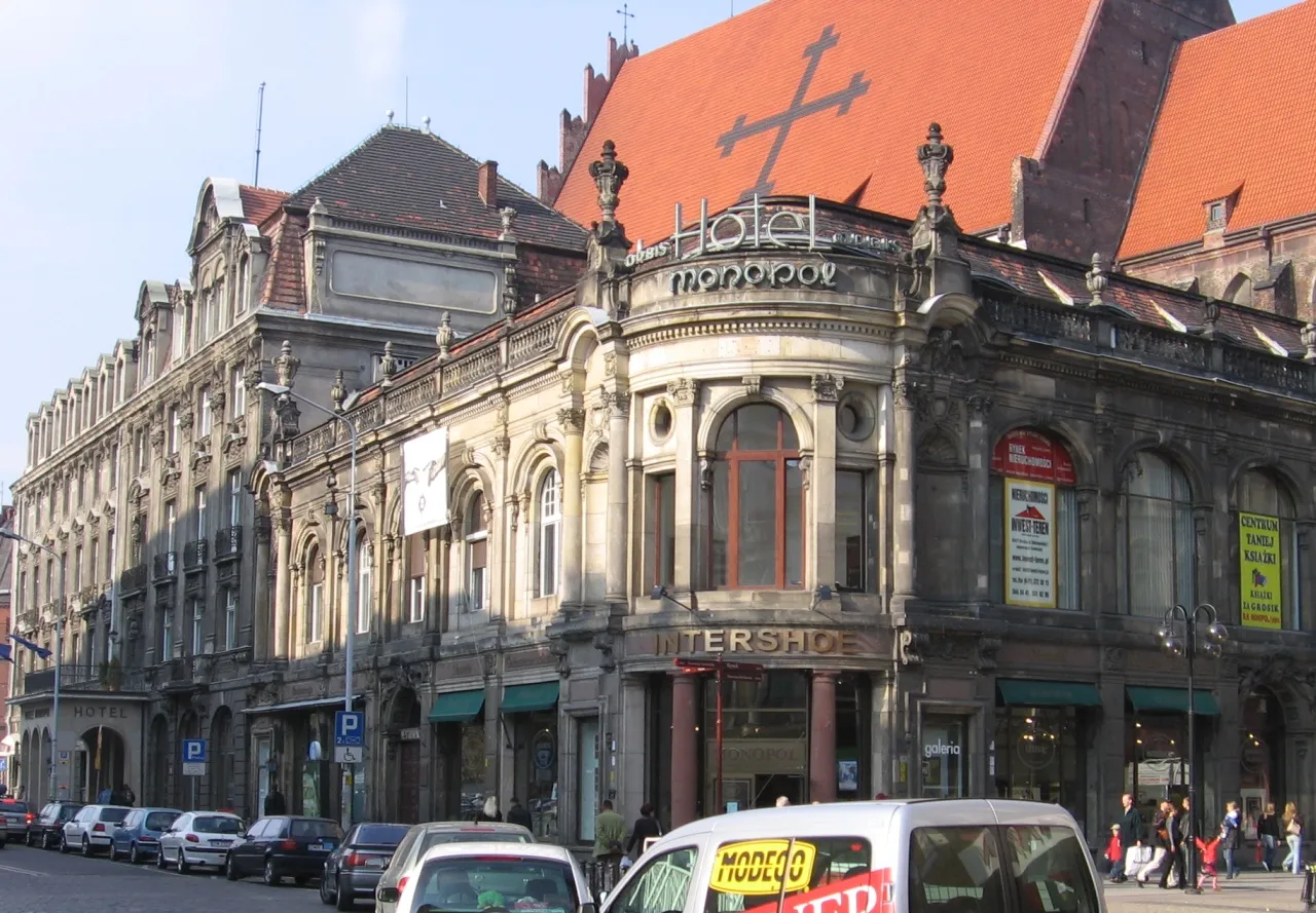 Narożnik Hotelu Monopol przed remontem: pociemniała elewacja, szyld Orbis i reklamy sklepów; samochody zaparkowane wzdłuż ulicy.